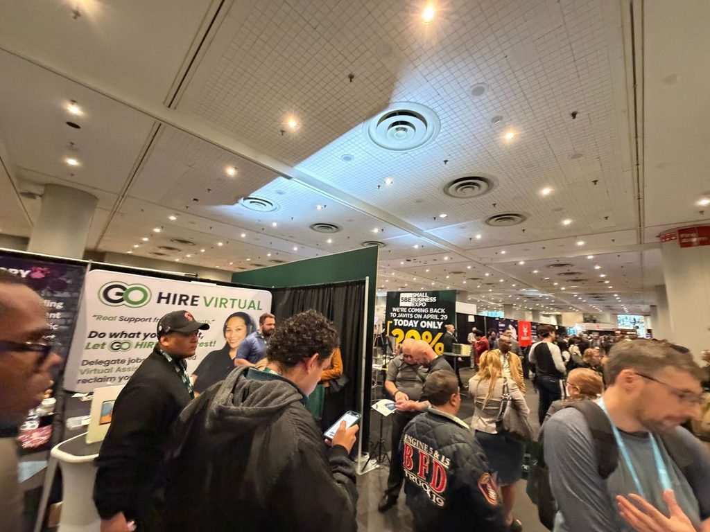 attendees-visiting-go-hire-virtual-booth-javits-center | Go Hire Virtual - Virtual Assistants for just Busy convention hall at a career fair; GO HIRE VIRTUAL booth with banner on the left and attendees walking nearby, many booths in the background.