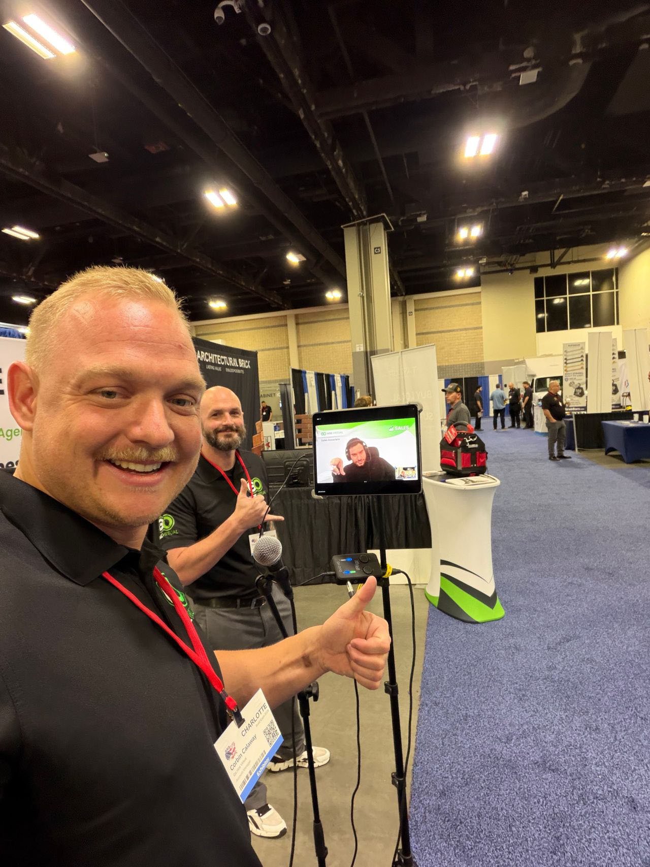 Smiling man in a black shirt takes a selfie at a convention booth; a tablet on a stand shows a video call while a second man stands behind with a microphone.