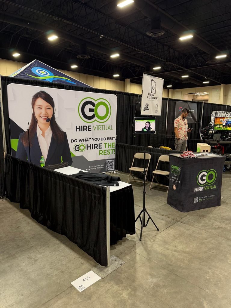 GO Hire Virtual trade show booth with a large banner showing a smiling woman in a suit and headset, black draped barriers, and a tablet display promoting virtual assistants at an expo.